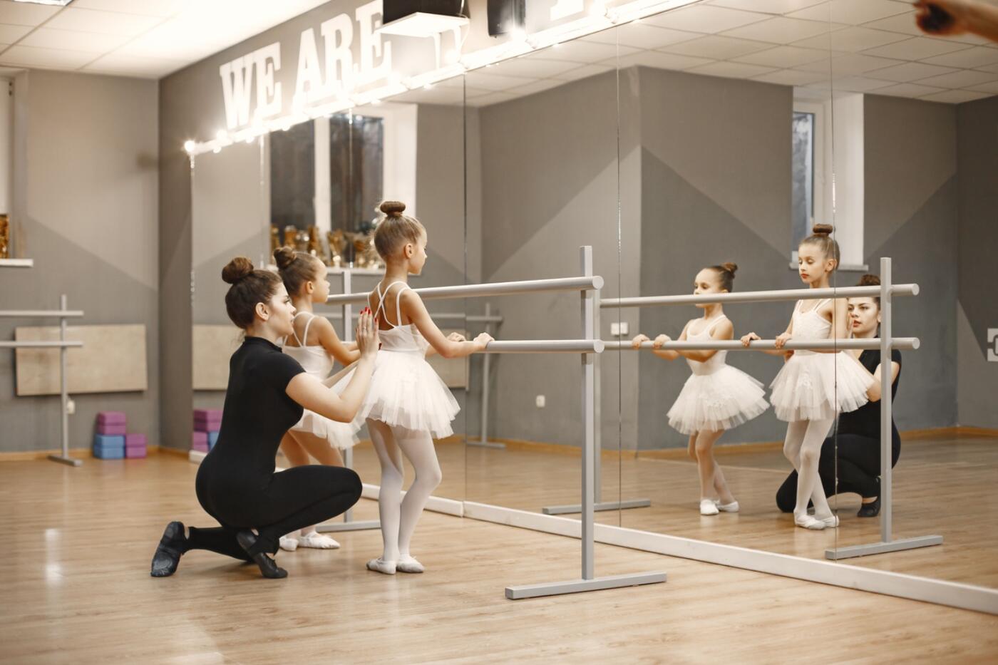 A teacher kneeling beside two young ballet students at the barre in a bright studio