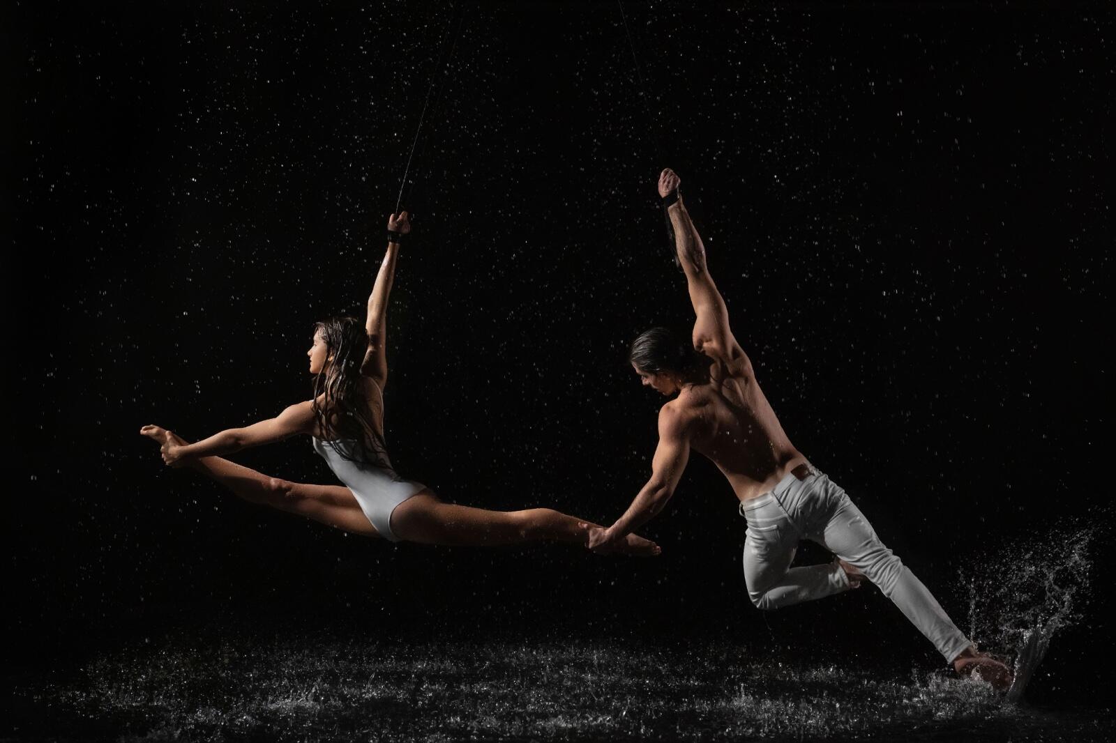 Two aerial dancers suspended mid-flight against a black stage, rain catching the light